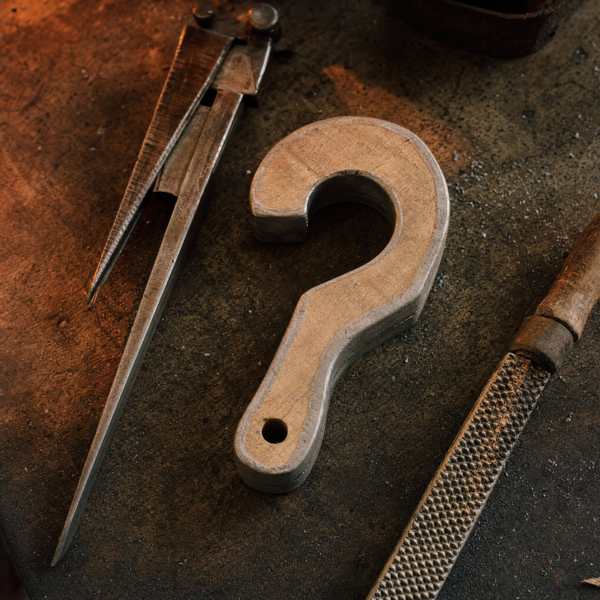 A wooden foundry pattern shaped like a wall hook, on a workbench with calipers and a rasp